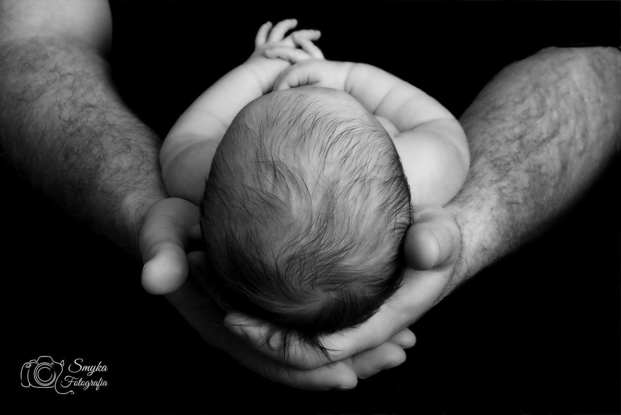 Czarno-białe zbliżenie główki noworodka w dłoniach dorosłego mężczyzny, z widocznymi włosami dziecka i fragmentami ramion ojca na czarnym tle.