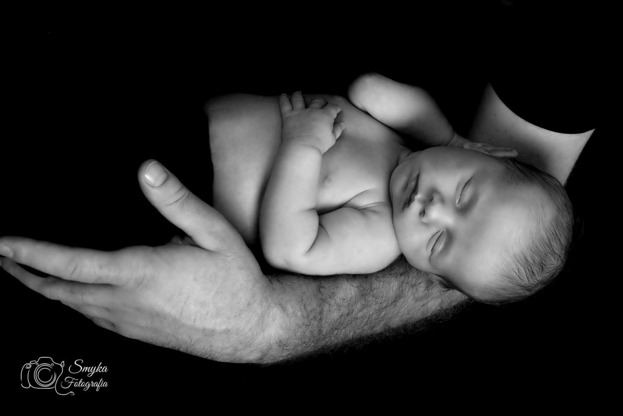 Czarno-białe zdjęcie noworodka śpiącego bezpiecznie w dłoni dorosłego, z widocznym logo fotografa w lewym dolnym rogu.