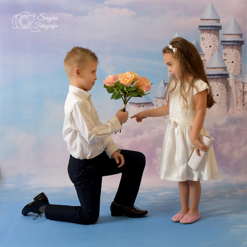 Chłopiec klęczy z bukietem róż, dziewczynka wskazuje na kwiaty, tło z motywem zamku i nieba.