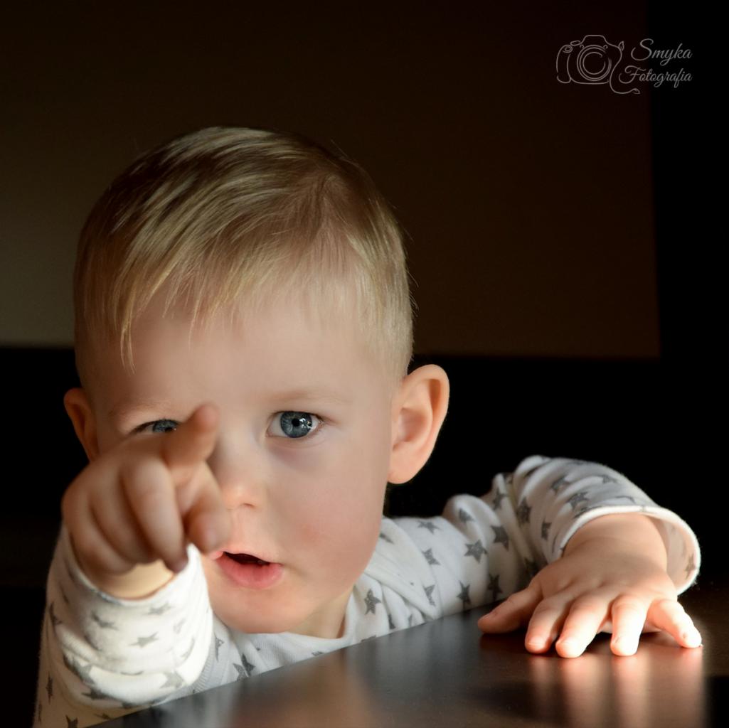 Portret blondwłosego chłopca z niebieskimi oczami, wskazującego palcem w kierunku widza, ubrany w białe body w szare gwiazdki, z logo fotografa w prawym górnym rogu.