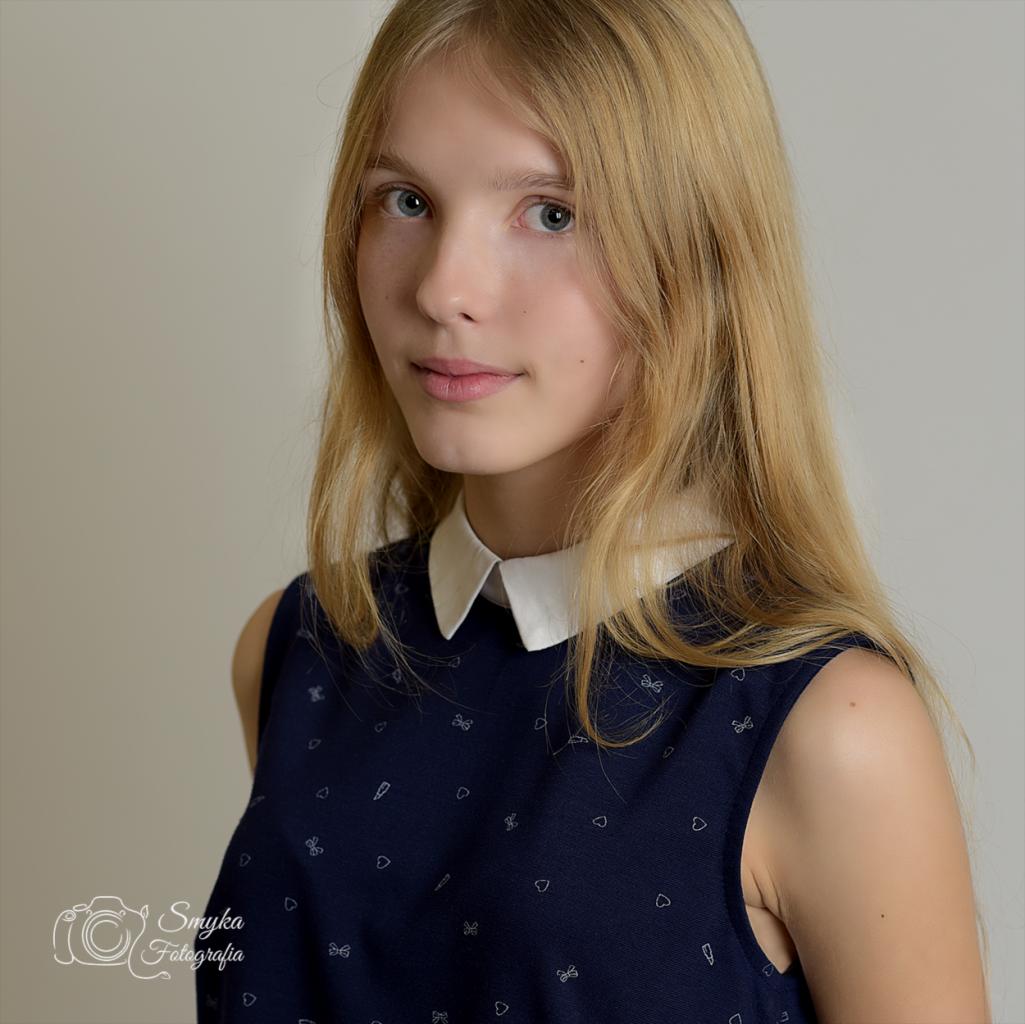 Portret młodej blondynki w granatowej sukience z białym kołnierzykiem, patrzącej w obiektyw na neutralnym tle.