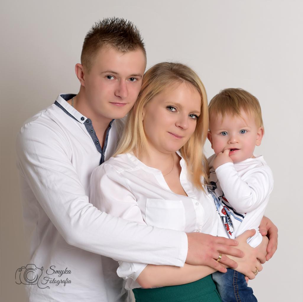 Portret rodziny: ojciec z modną fryzurą obejmuje matkę trzymającą synka z palcem w buzi, wszyscy ubrani w białe koszule na jasnym tle.