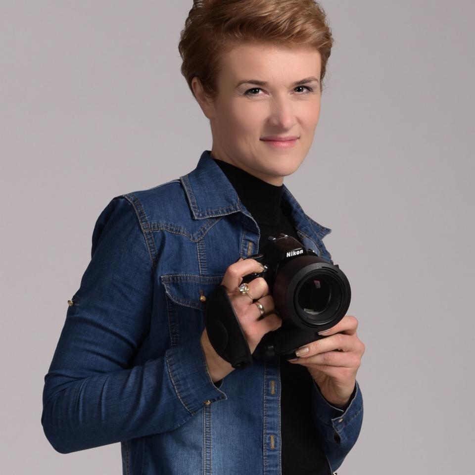 Portret uśmiechniętej kobiety z krótkimi włosami w dżinsowej koszuli, trzymającej aparat Nikon z dużym obiektywem i pierścionkami na palcach.