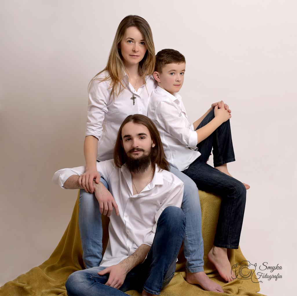 Portret rodzinny: matka, ojciec i syn w białych koszulach i jeansach, pozujący na żółtym tle w studio, widoczne bose stopy chłopca i ojca.