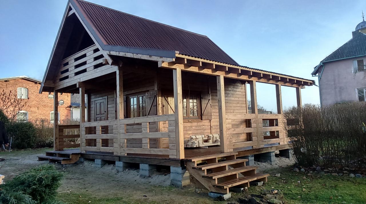 Drewniany dom z bali z brązowym dachem z blachy falistej, w trakcie budowy, z widocznymi fundamentami z betonowych bloczków i schodami wejściowymi, na tle innych budynków i zieleni.
