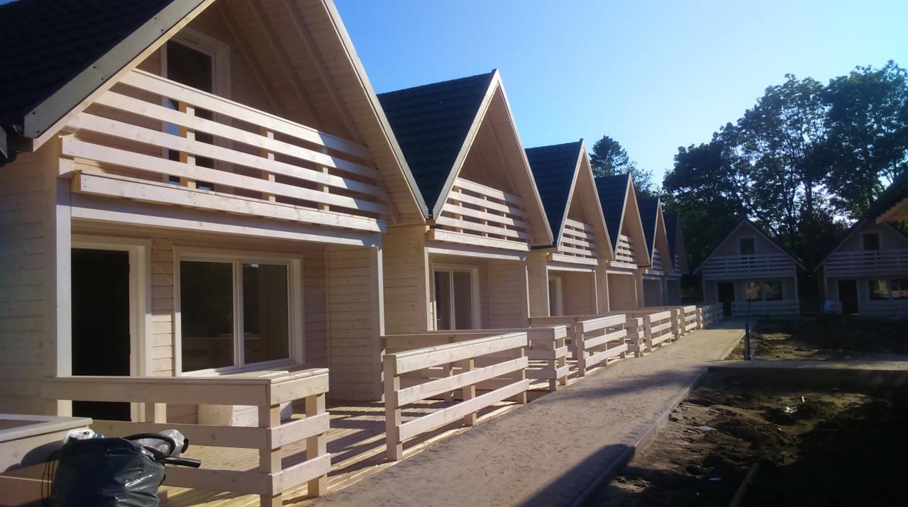 Szereg nowo wybudowanych, identycznych domków letniskowych z jasnego drewna, z balkonami i ciemnymi dachami, ustawionych w rzędzie w słoneczny dzień.