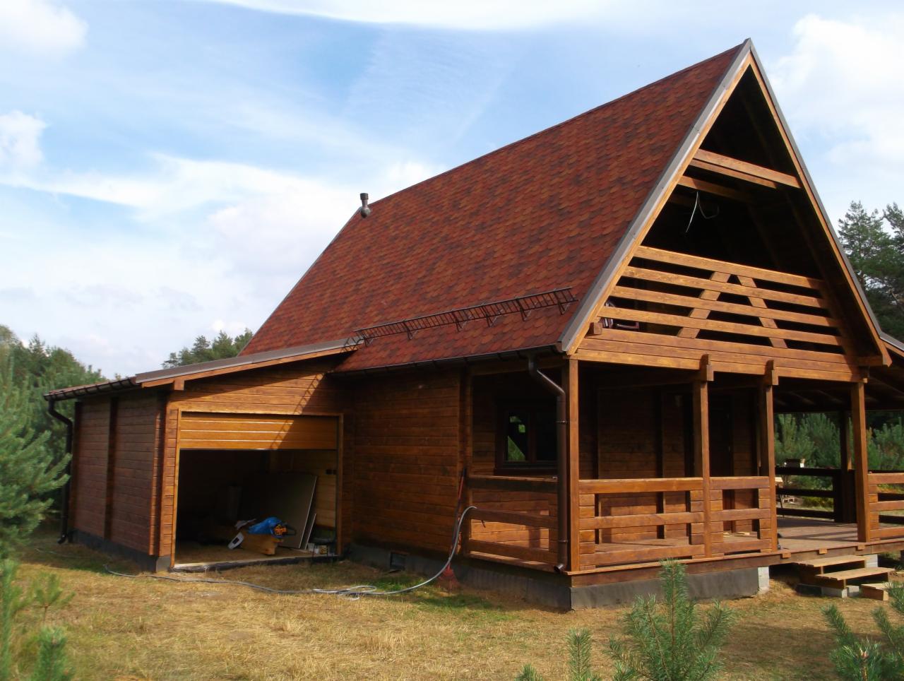 Surowy, drewniany dom w trakcie budowy z otwartym garażem, widoczne narzędzia w środku, brązowy dach z gontu bitumicznego, otoczenie leśne.