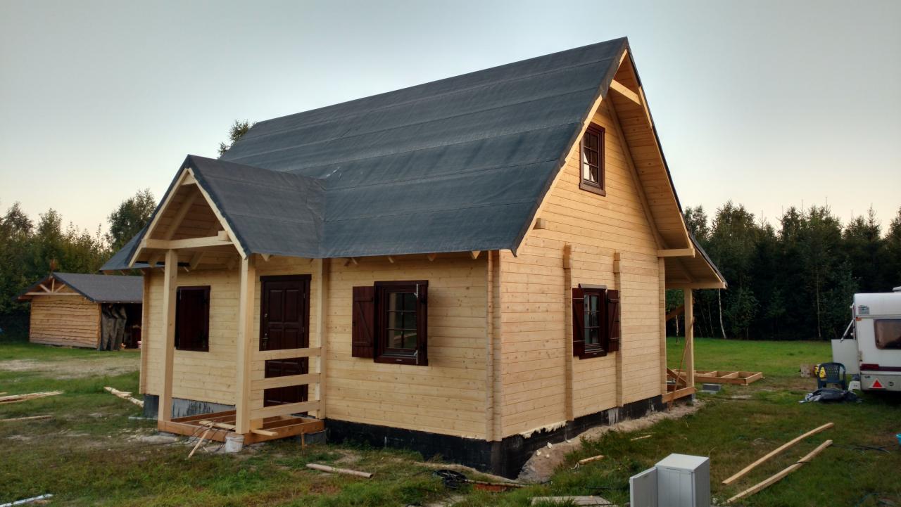Surowy, drewniany dom z bali z ciemnymi okiennicami i dachem pokrytym papą, w trakcie budowy, widok z zewnątrz, na trawiastym terenie z drzewami w tle.