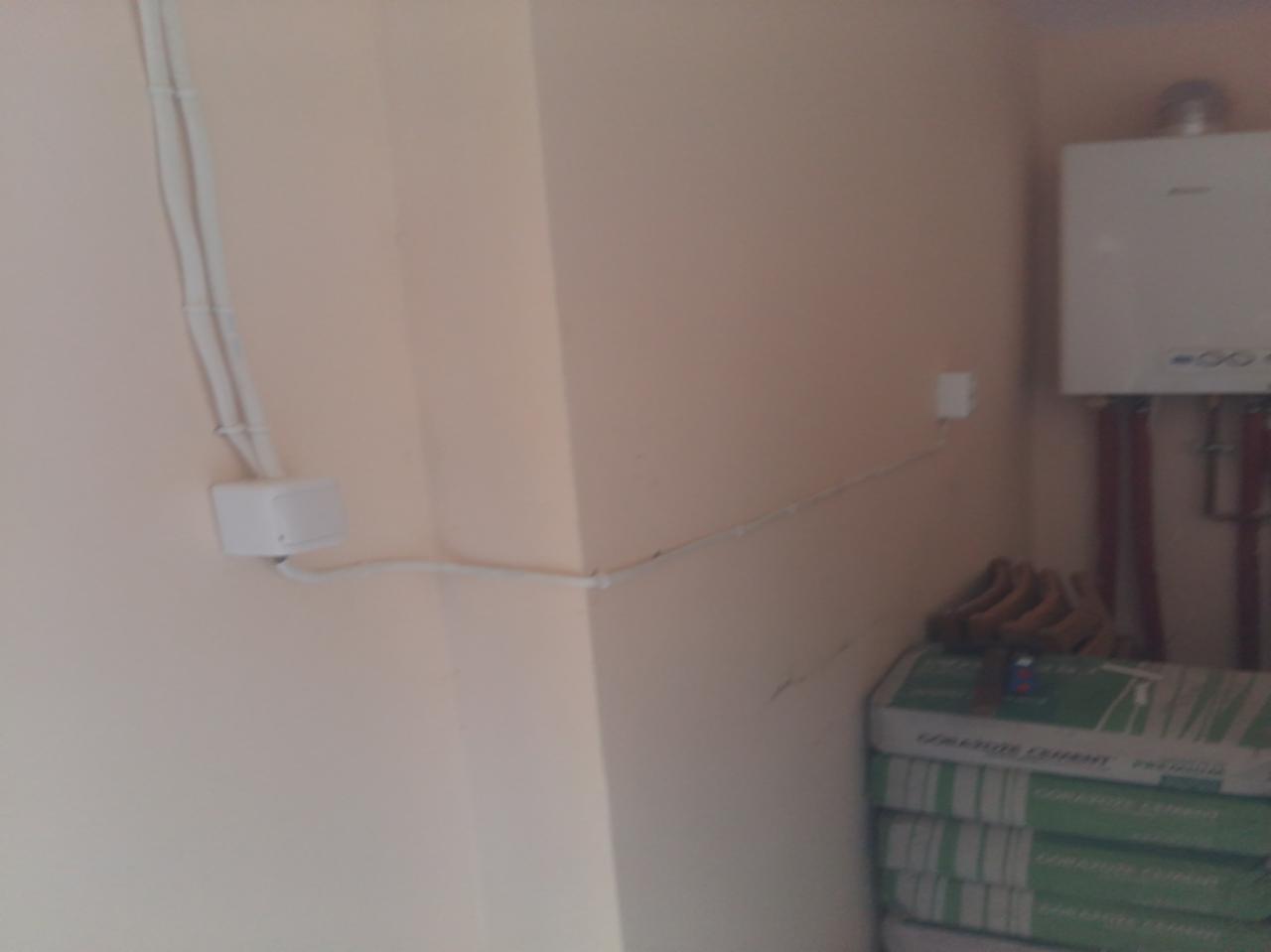 Biała puszka elektryczna z kablami na beżowej ścianie, obok piec gazowy i worki z cementem