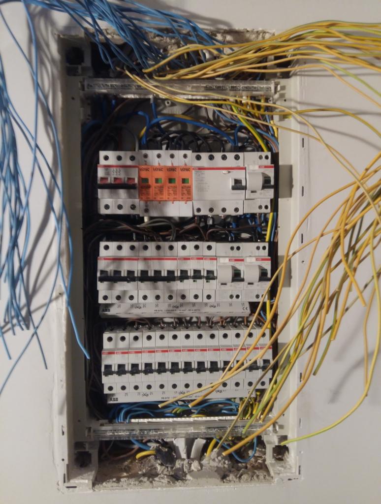 Otwarta skrzynka rozdzielcza z widocznymi zabezpieczeniami, przewodami w kolorach niebieskim i żółtym, oraz elementami montażowymi w ścianie.