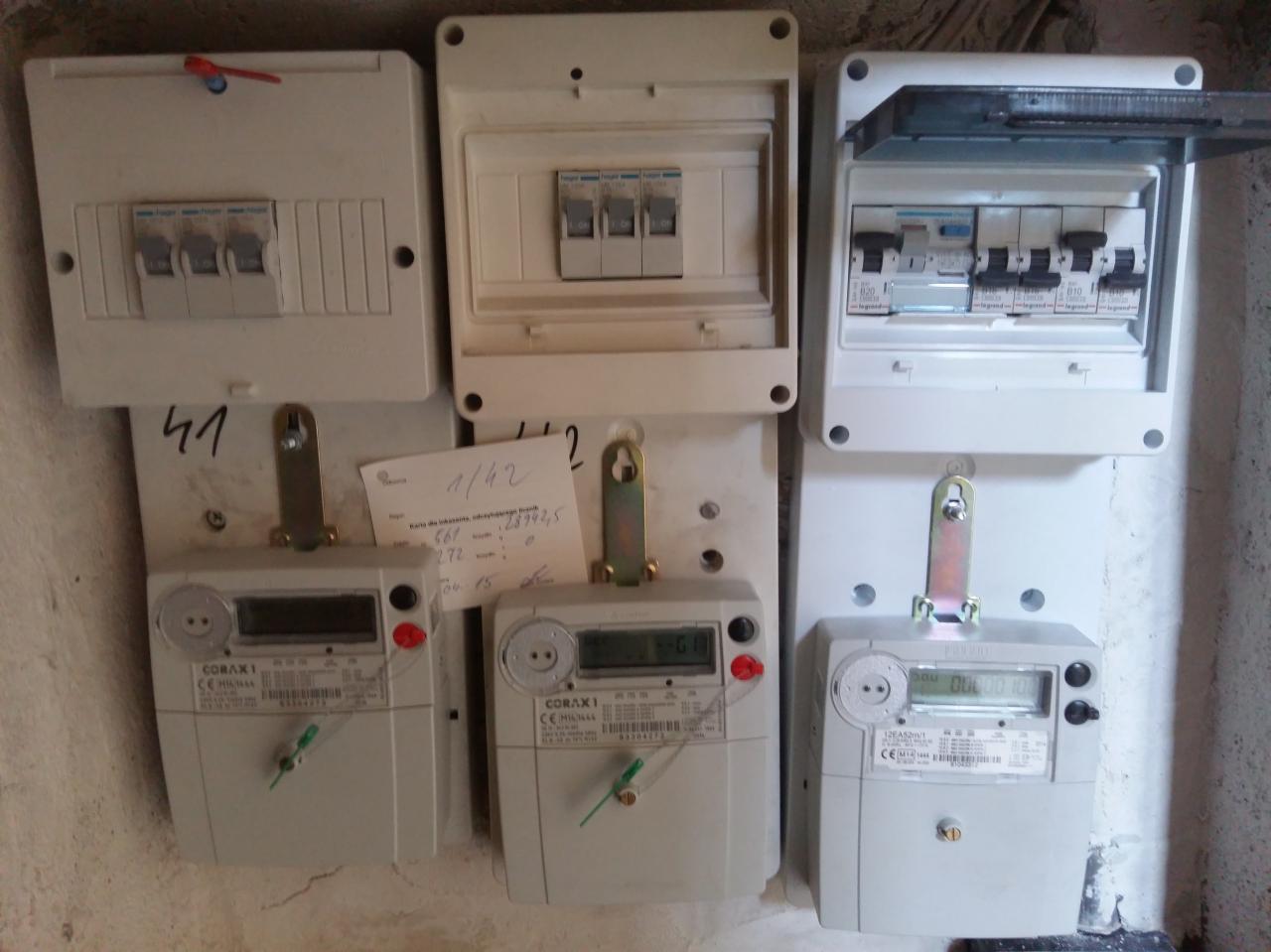 Trzy elektryczne skrzynki rozdzielcze i trzy liczniki zużycia energii elektrycznej zamontowane obok siebie na ścianie. Na jednym z liczników przyczepiona kartka z odczytem.