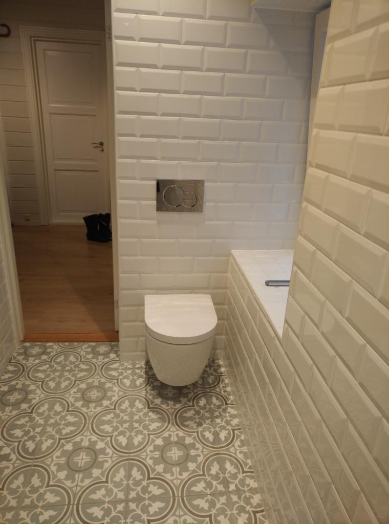 Biała toaleta wisząca na tle ściany wyłożonej białymi, wypukłymi płytkami typu metro, z widocznym chromowanym przyciskiem spłuczki i podłogą z szarych płytek w geometryczne wzory.