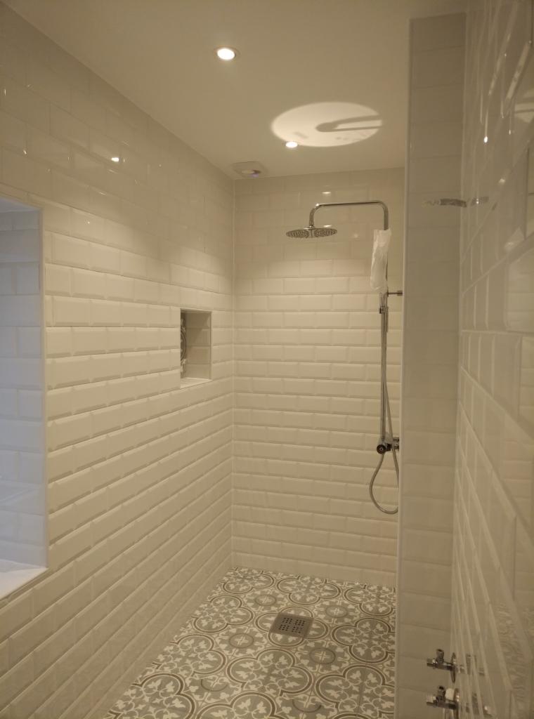 Nowoczesna kabina prysznicowa z białymi, prostokątnymi płytkami ułożonymi w cegiełkę, wzorzysta szaro-biała podłoga i wnęka w ścianie.