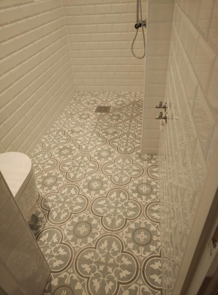 Wykończona łazienka z białą armaturą, ściany wyłożone białymi, prostokątnymi płytkami, podłoga z płytek w szaro-białe, kwieciste wzory, widoczny odpływ liniowy.