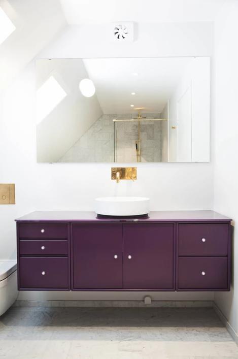 Nowoczesna łazienka z fioletową szafką pod umywalką, okrągłą białą umywalką nablatową, złotą baterią i dużym lustrem, w którym odbija się kabina prysznicowa z elementami w kolorze złota.