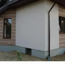 Fragment domu z otynkowaną elewacją w kolorze kremowym, z brązowymi elementami dekoracyjnymi i oknami, rynna pionowa, widoczny fragment fundamentu i otoczenie posesji w trakcie prac wykończeniowych.