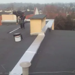 Prace dekarskie na dachu pokrytym papą termozgrzewalną, widoczny pracownik w uprzęży, zabezpieczenia komina blachą, puszka z impregnatem do papy.