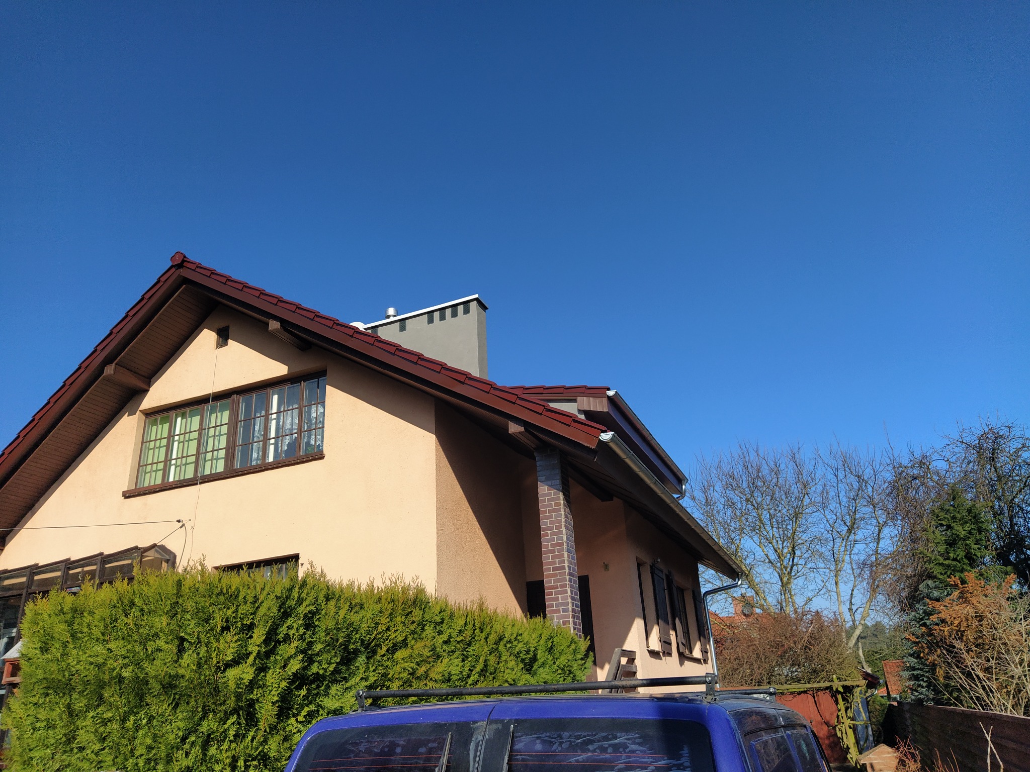 Dom jednorodzinny z dwuspadowym dachem pokrytym czerwoną dachówką, widok z dołu na tle błękitnego nieba i częściowo zasłonięty przez żywopłot i samochód.