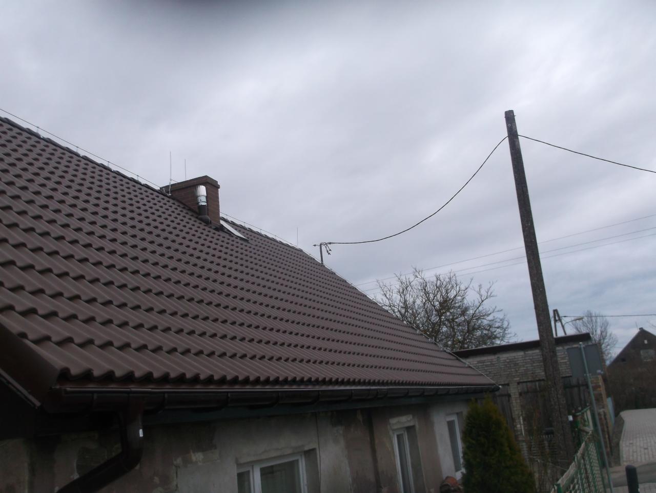 Brązowy dach z dachówki ceramicznej na tle pochmurnego nieba, widoczny komin i rynna, słup energetyczny w tle.