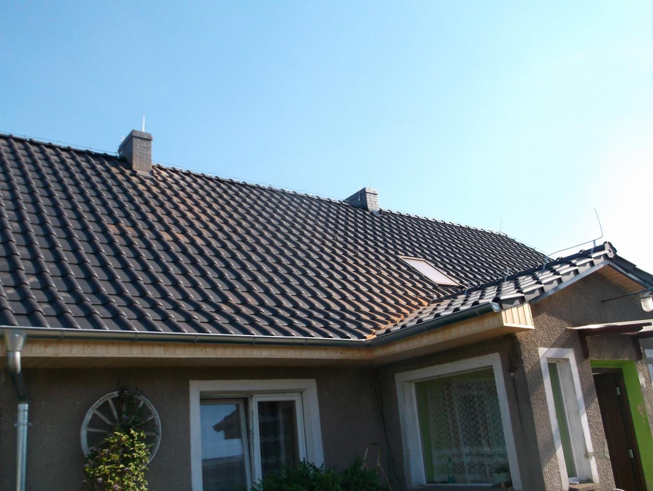 Dom jednorodzinny z ciemną dachówką ceramiczną i oknem dachowym, widok z dołu na tle błękitnego nieba, widoczne rynny i fragment elewacji z dekoracyjnym kołem.