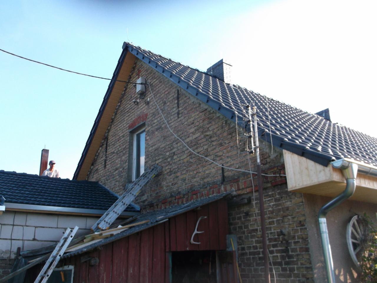 Dwuspadowy dach z ciemnej dachówki ceramicznej na murowanym budynku, widoczna drabina oparta o ścianę, rynna i antena, pracownik na dachu sąsiedniego budynku.
