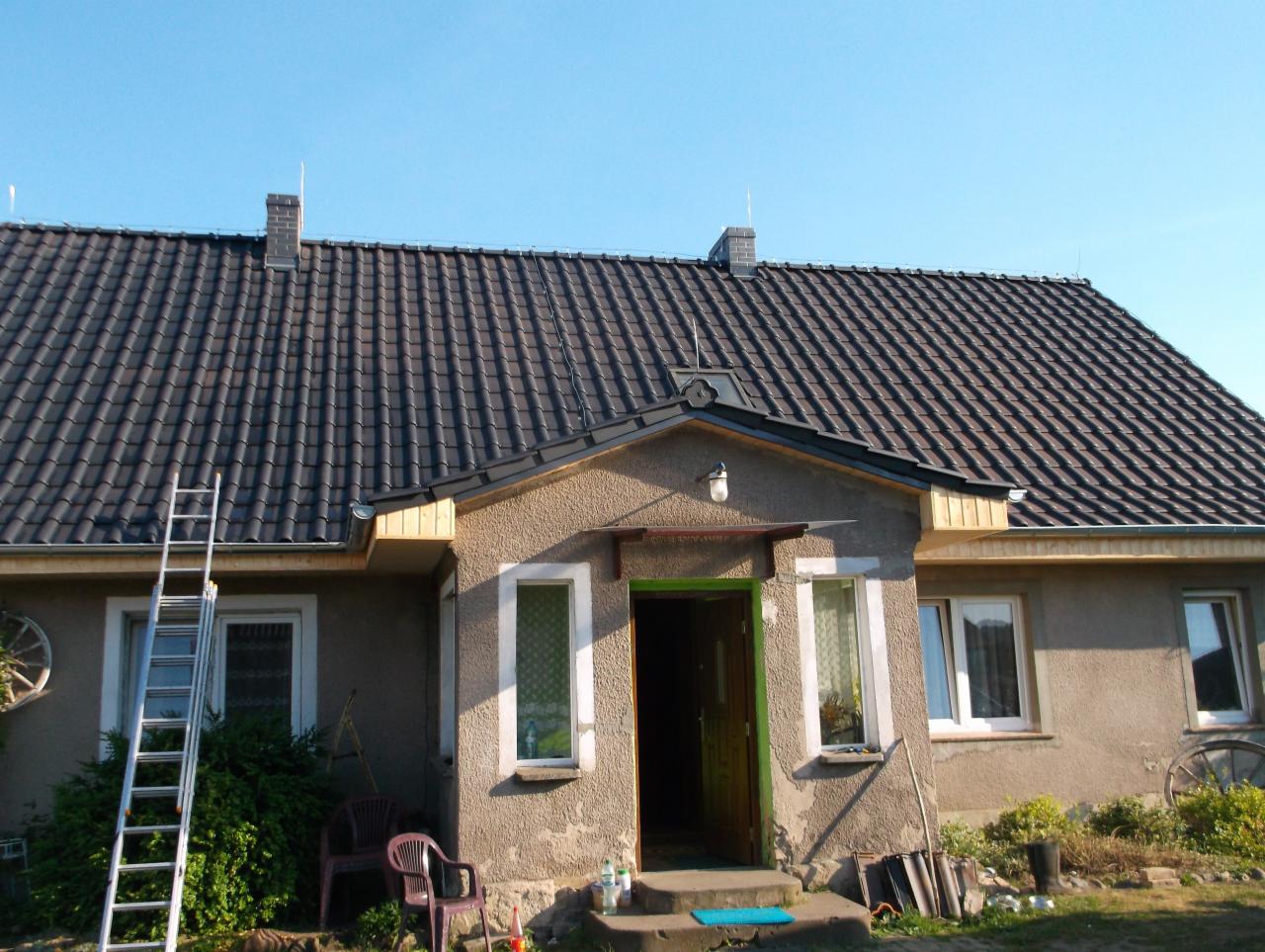 Dom jednorodzinny z ciemną dachówką karpiówką, widoczna drabina oparta o ścianę, teren posesji częściowo zagospodarowany.