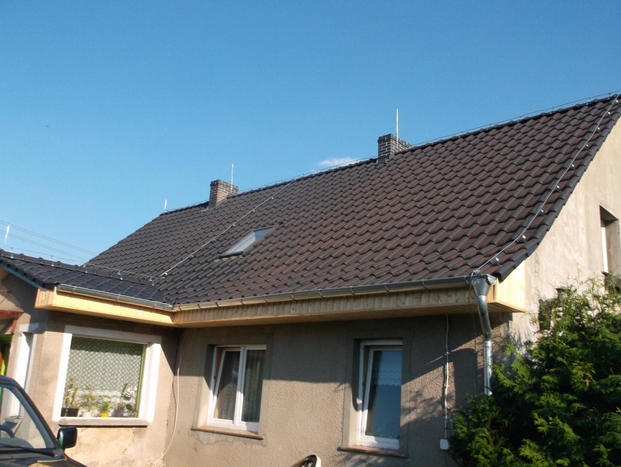 Dom z nowym, ciemnobrązowym dachem z dachówki ceramicznej, widoczne okno dachowe, komin i nowo zamontowane rynny, fragment samochodu w lewym dolnym rogu.