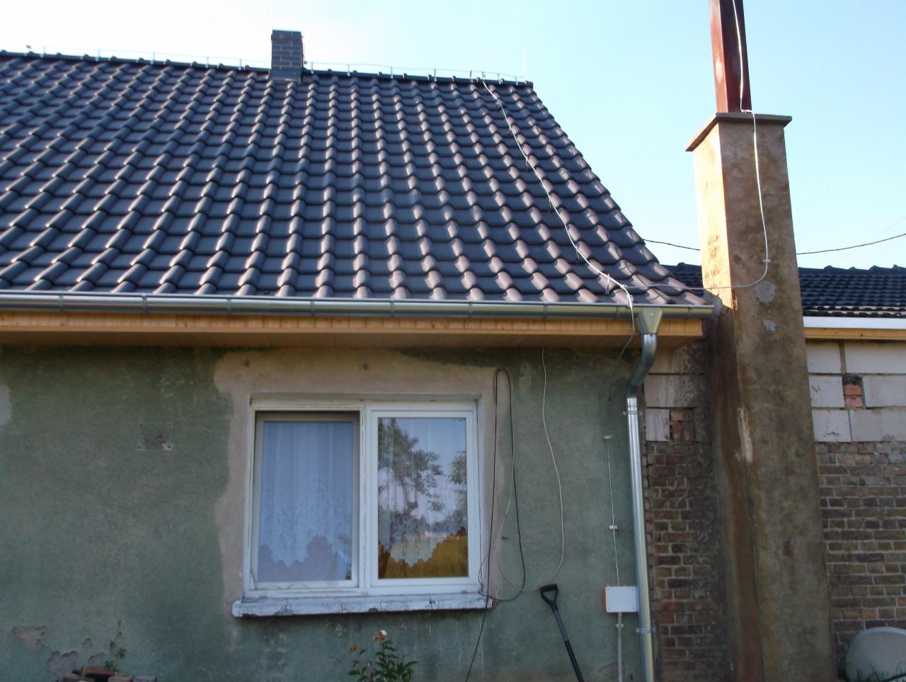 Dom z nowym, ciemnoszarym dachem z dachówki ceramicznej, widoczne rynny i komin, elewacja w trakcie naprawy, okno z firanką.