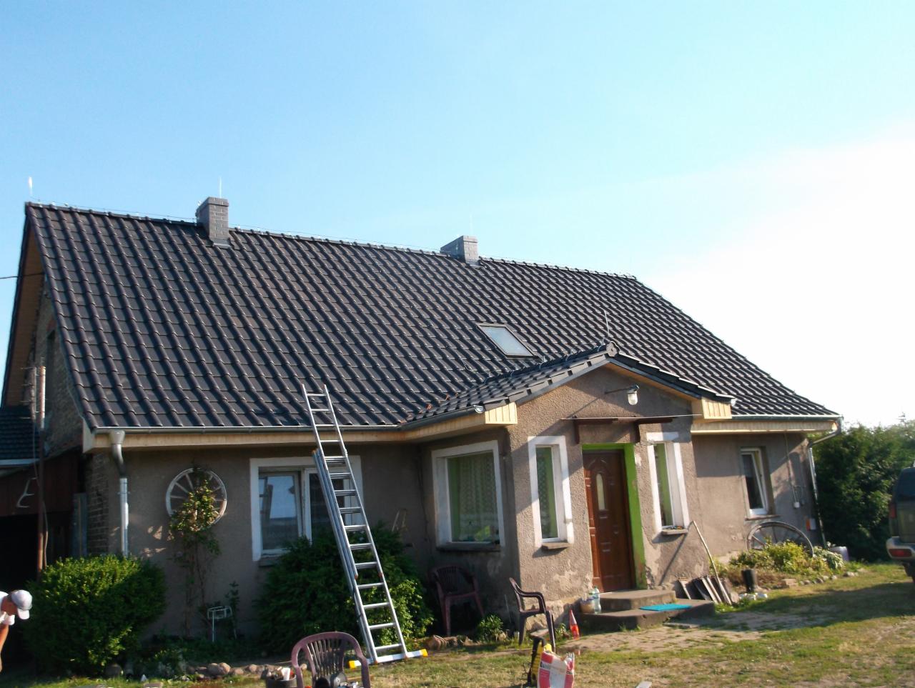 Dom jednorodzinny z nowym, ciemnoszarym dachem z dachówki ceramicznej, widoczna drabina oparta o ścianę, dwa kominy i okno dachowe.