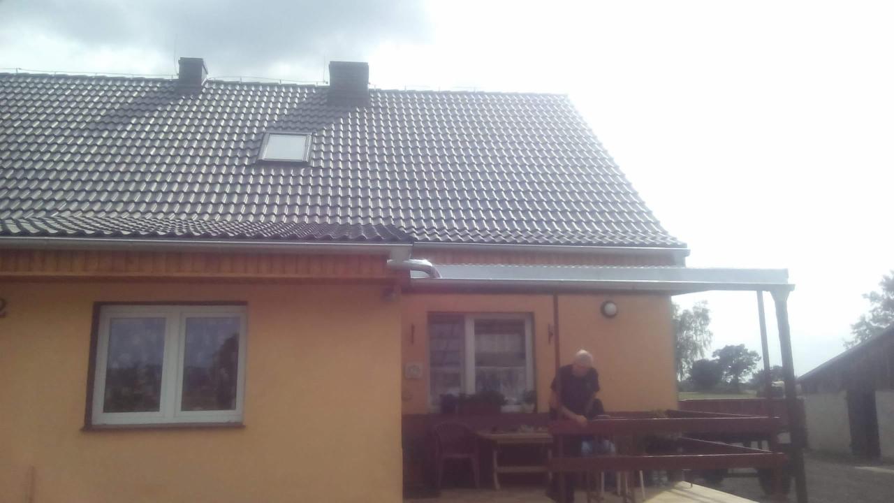 Dom jednorodzinny z ciemną dachówką, widoczne okno dachowe i orynnowanie, fragment tarasu z osobą w tle. Jasne niebo, lekko prześwietlone zdjęcie.