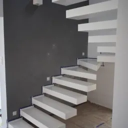 Nowoczesne, białe schody wspornikowe z mikrocementową szarą ścianą w tle, zabezpieczone folią malarską podczas prac wykończeniowych.