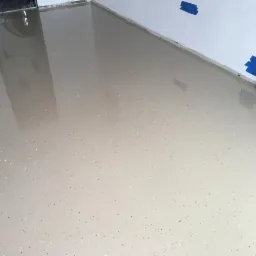 Jasnoszara, błyszcząca posadzka z delikatnym, wielobarwnym wzorem punktowym, zabezpieczona niebieską taśmą malarską przy ścianie i rurami instalacyjnymi.