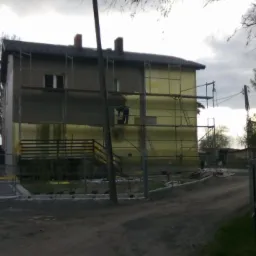 Dwukondygnacyjny dom w trakcie prac ociepleniowych, z widocznymi rusztowaniami i częściowo nałożonym styropianem na elewacji, zarys postaci pracownika na rusztowaniu