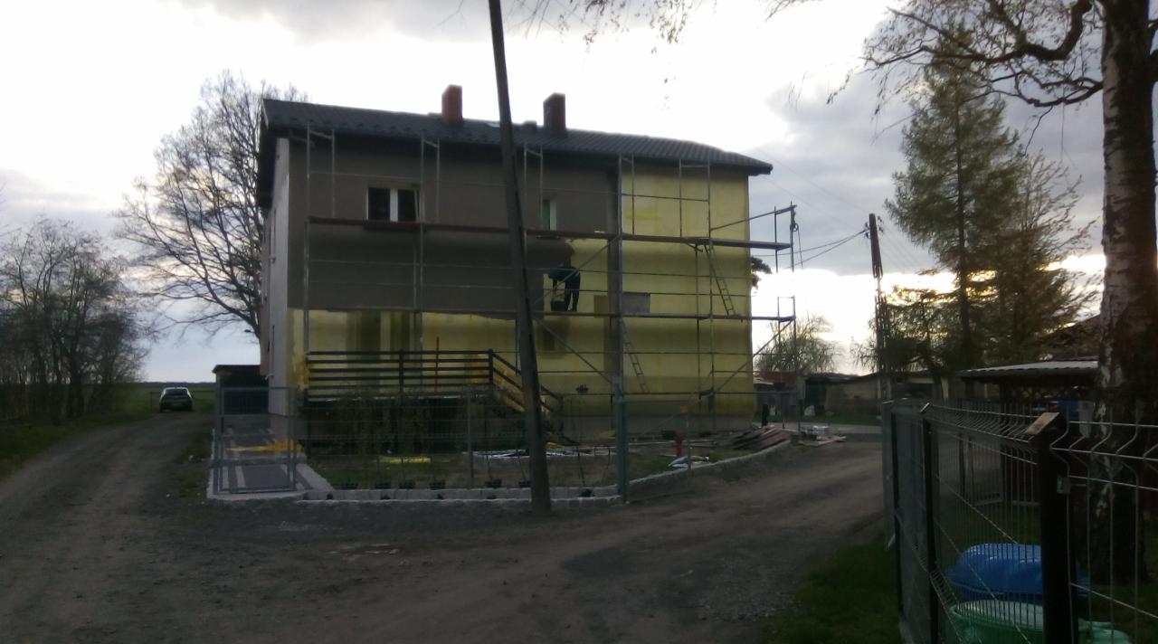 Dwukondygnacyjny dom w trakcie prac ociepleniowych, z widocznymi rusztowaniami i częściowo nałożonym styropianem na elewacji, zarys postaci pracownika na rusztowaniu