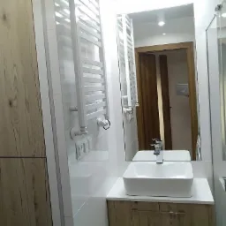 Nowoczesna łazienka z drewnopodobnymi szafkami, białymi płytkami, grzejnikiem drabinkowym i prostokątną umywalką nablatową; w lustrze odbija się fragment drzwi i osoba robiąca zdjęcie.