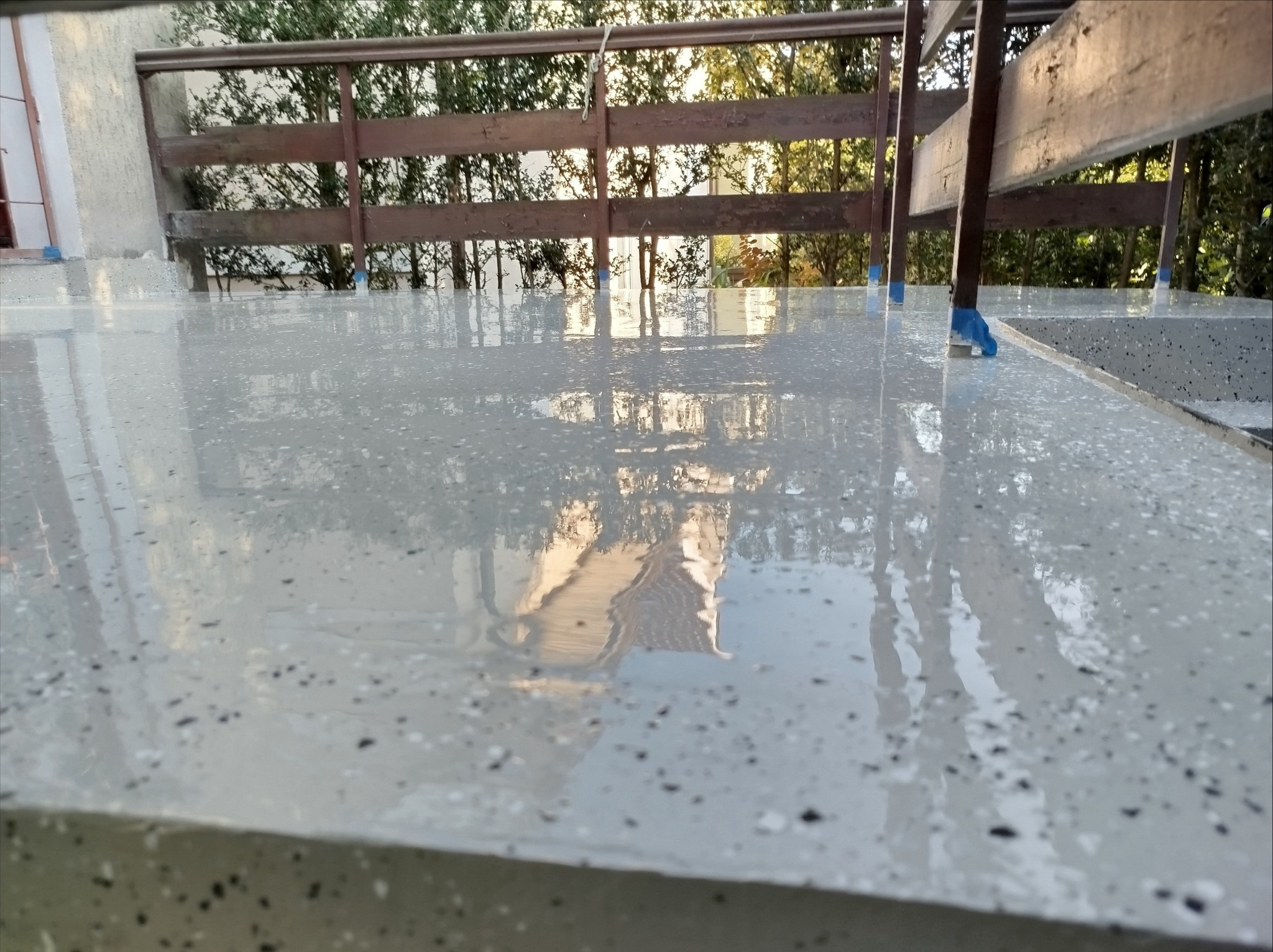 Świeżo wylana, jasnoszara posadzka żywiczna z ciemnymi plamkami na tarasie biurowym, zabezpieczona niebieską taśmą malarską przy drewnianej balustradzie.