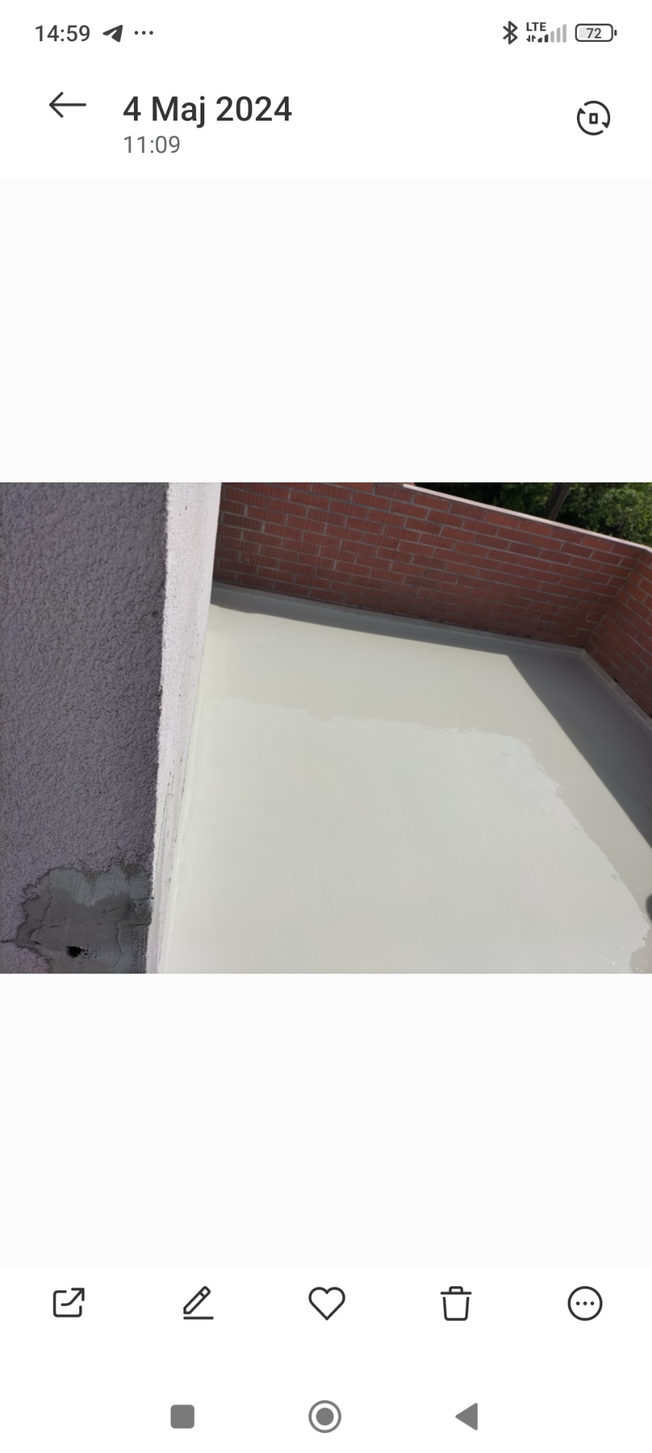 Biała, gładka powierzchnia nowej wylewki na tarasie lub balkonie, z widoczną ścianą z czerwonej cegły w tle i fragmentem otynkowanej ściany z widocznymi śladami naprawy po lewej stronie.