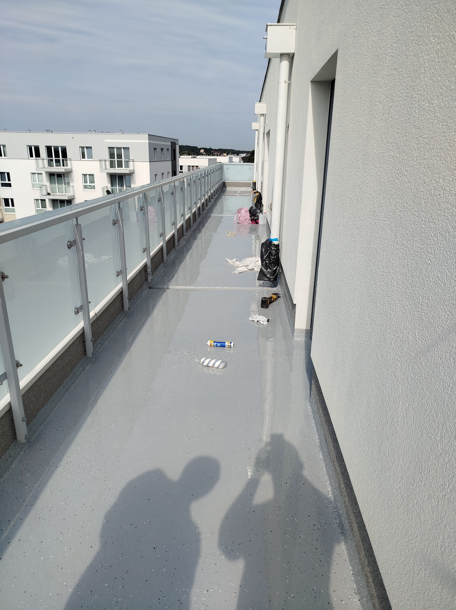 Szary balkon z żywicy epoksydowej po remoncie, widoczne narzędzia malarskie, śmieci i cień osoby robiącej zdjęcie.