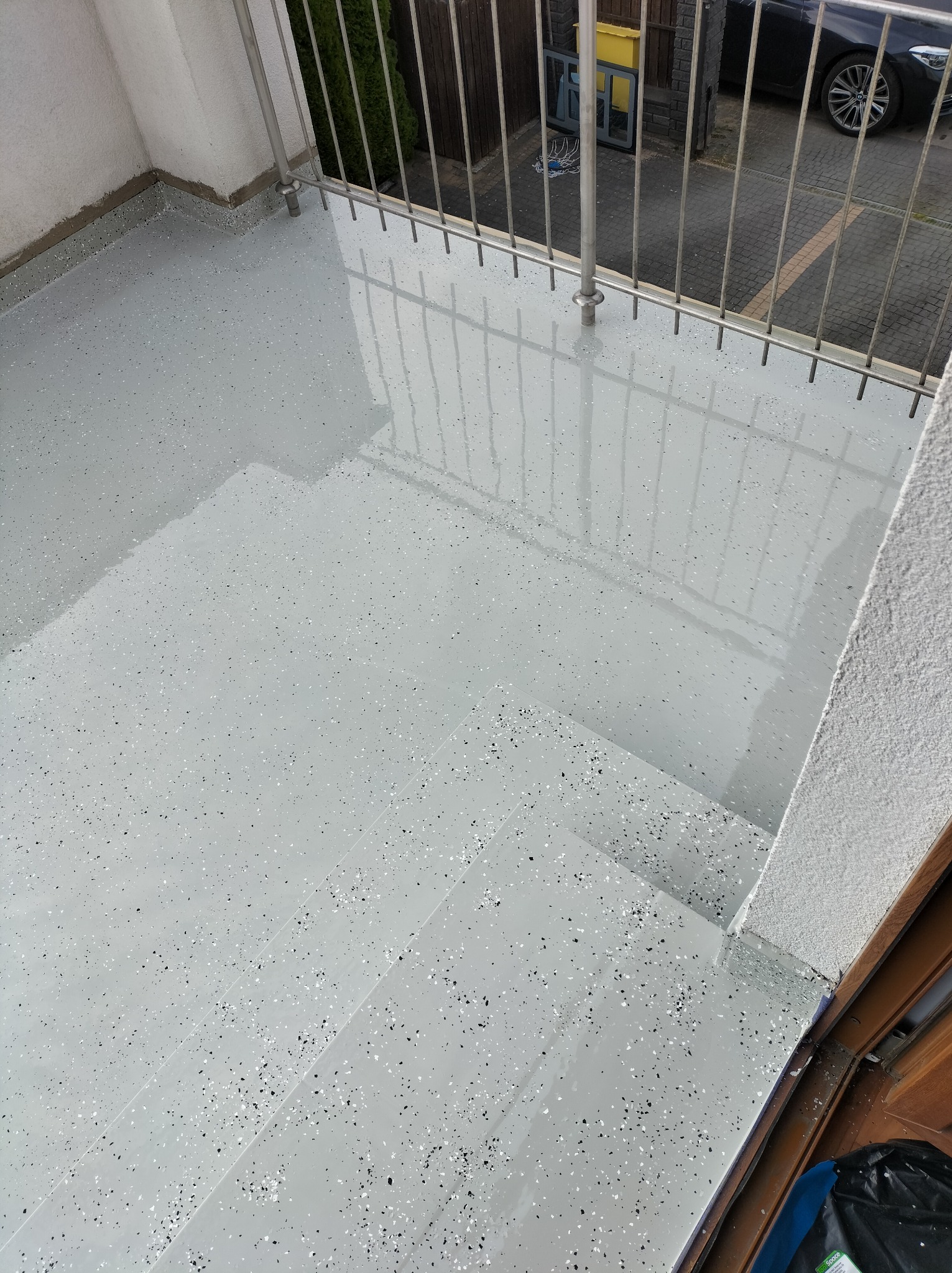 Świeżo wylana, jasnoszara posadzka żywiczna z czarnymi i białymi drobinami na balkonie, widoczne schody i metalowa balustrada, odbijająca światło.