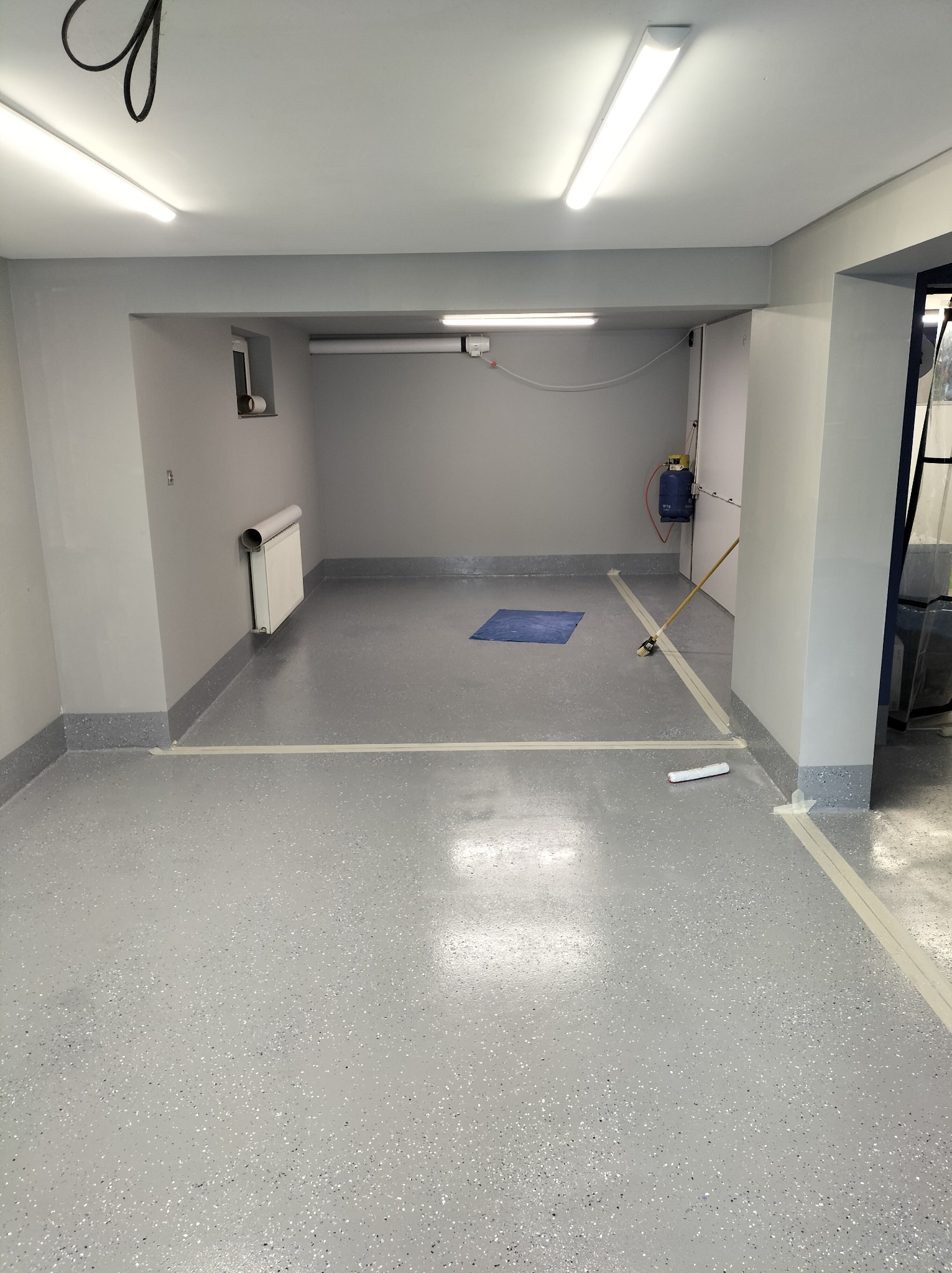 Szara, przemysłowa posadzka żywiczna z kolorowymi drobinkami w świeżo odremontowanym garażu lub pomieszczeniu biurowym, zabezpieczona taśmą malarską przy ścianach, z widocznym grzejnikiem...