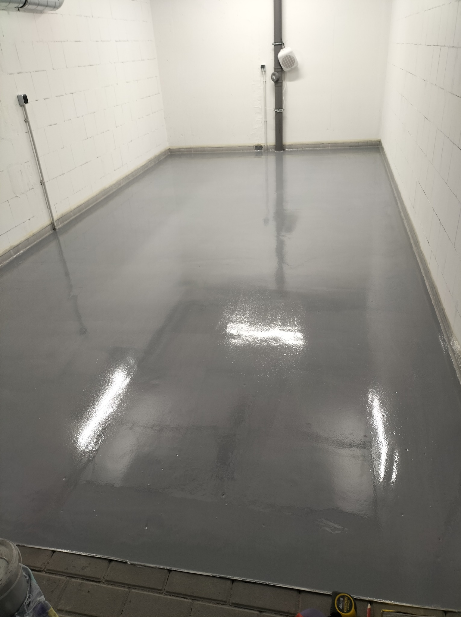 Wykończona szara posadzka żywiczna w pomieszczeniu biurowym z białymi ścianami z betonu komórkowego, widoczna rura kanalizacyjna przy suficie i fragment taśmy mierniczej w rogu.