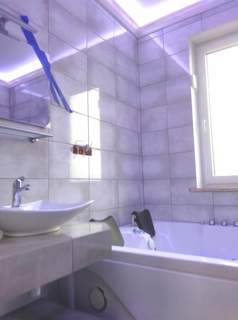 Łazienka w trakcie remontu: biała umywalka z baterią, wanna z hydromasażem, szare płytki ścienne, widoczne przewody elektryczne i niebieska taśma malarska.