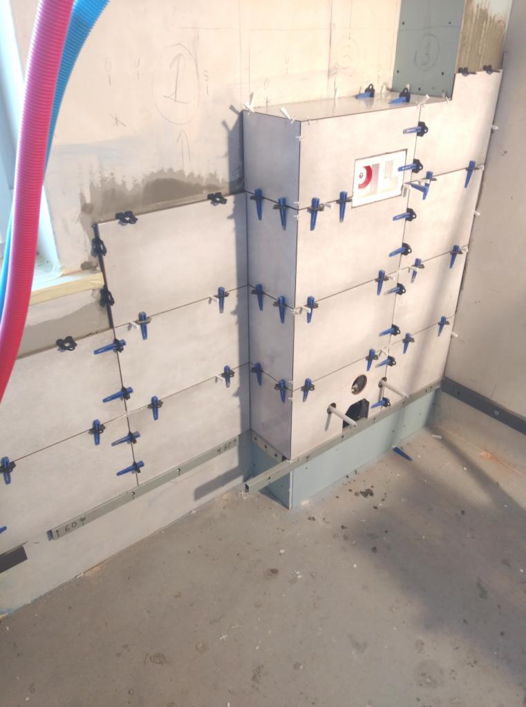 Świeżo ułożone jasnoszare płytki na ścianie, zabezpieczone krzyżykami dystansowymi, widoczne rury instalacji wodnej i fragment ściany z odręcznymi notatkami.