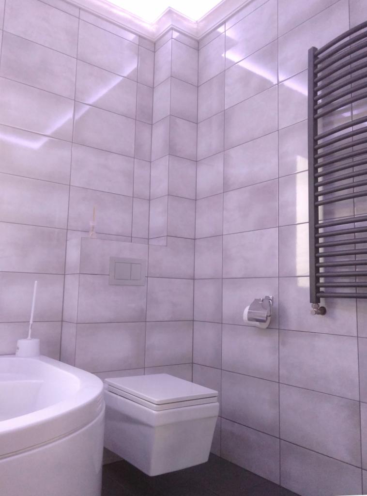 Wykończona łazienka z szarymi płytkami, wiszącą toaletą, grzejnikiem drabinkowym i podświetleniem LED przy suficie.