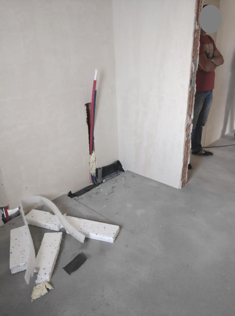 Przygotowanie do prac glazurniczych w rogu pomieszczenia: widoczne rury, styropianowe płyty izolacyjne, fragment posadzki oraz osoba w tle.