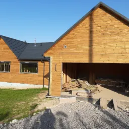 Surowy stan wykończenia zewnętrznego domu z drewnianą elewacją, widoczne materiały budowlane w otwartym garażu, teren przygotowany pod dalsze prace.