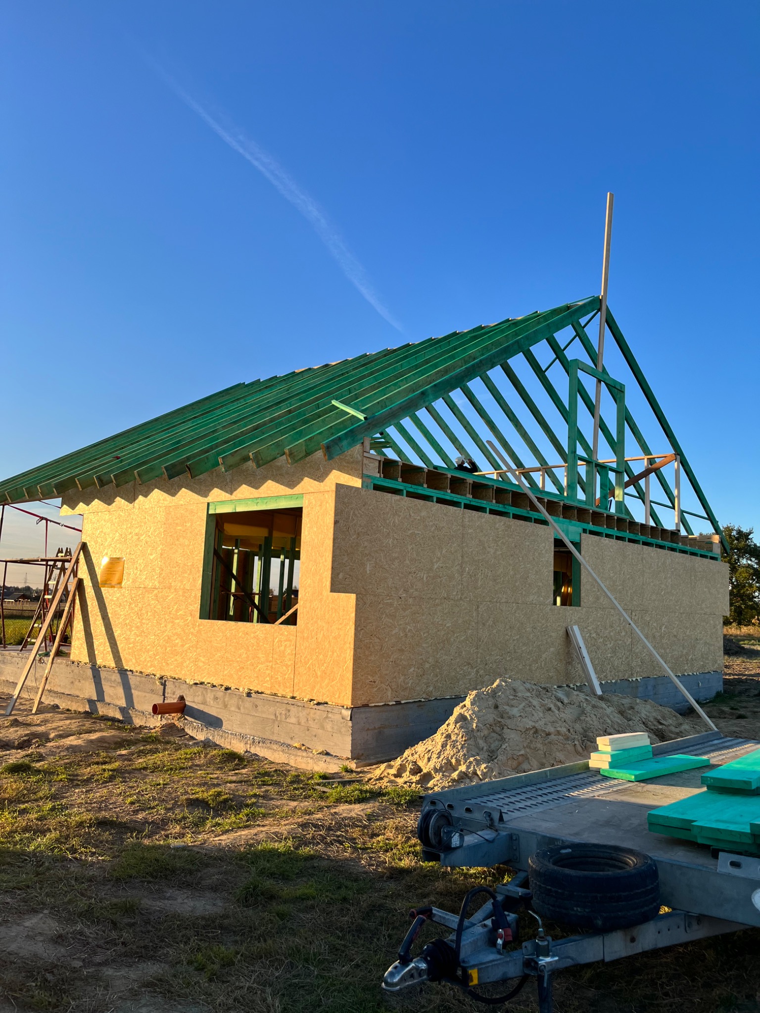 Wznoszenie budynku w technologii szkieletowej: surowe ściany z płyt OSB, zielona konstrukcja dachu w trakcie montażu, przyczepa z materiałami budowlanymi na pierwszym planie, błękitne niebo w tle.