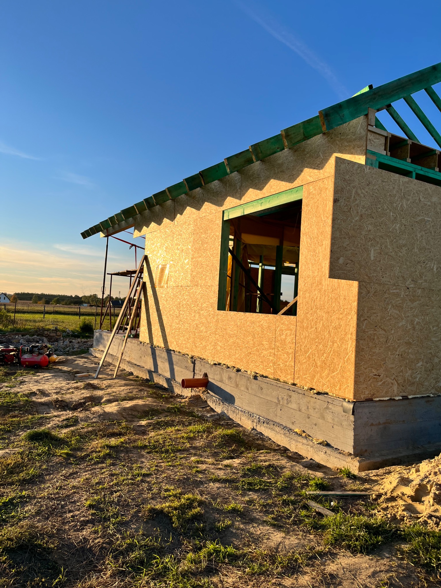 Wznoszenie konstrukcji budynku z drewnianych paneli OSB z widocznym szkieletem dachu, na fundamencie betonowym z rurą odpływową, w słoneczny dzień.