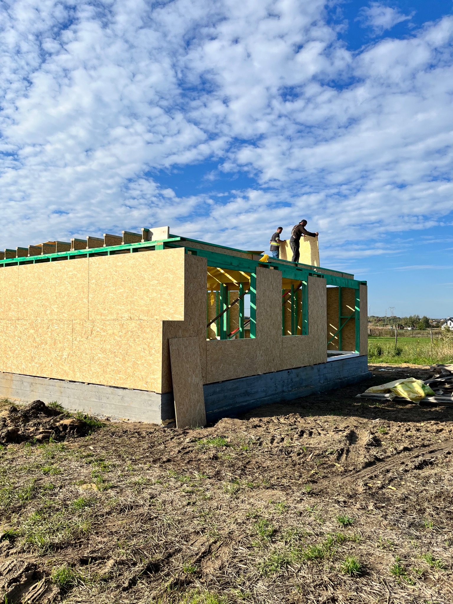 Montaż płyty OSB na ścianie zewnętrznej surowego stanu domu szkieletowego, widoczne zielone elementy konstrukcyjne, dwóch pracowników na dachu, błękitne niebo z chmurami w tle.