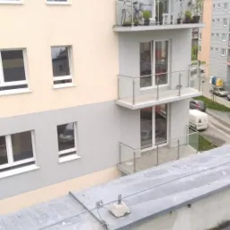 Wielorodzinny budynek po remoncie elewacji, widok z dachu, z widocznymi oknami i balkonami z szklanymi balustradami, na jednym z balkonów stoi rower i doniczki z roślinami.